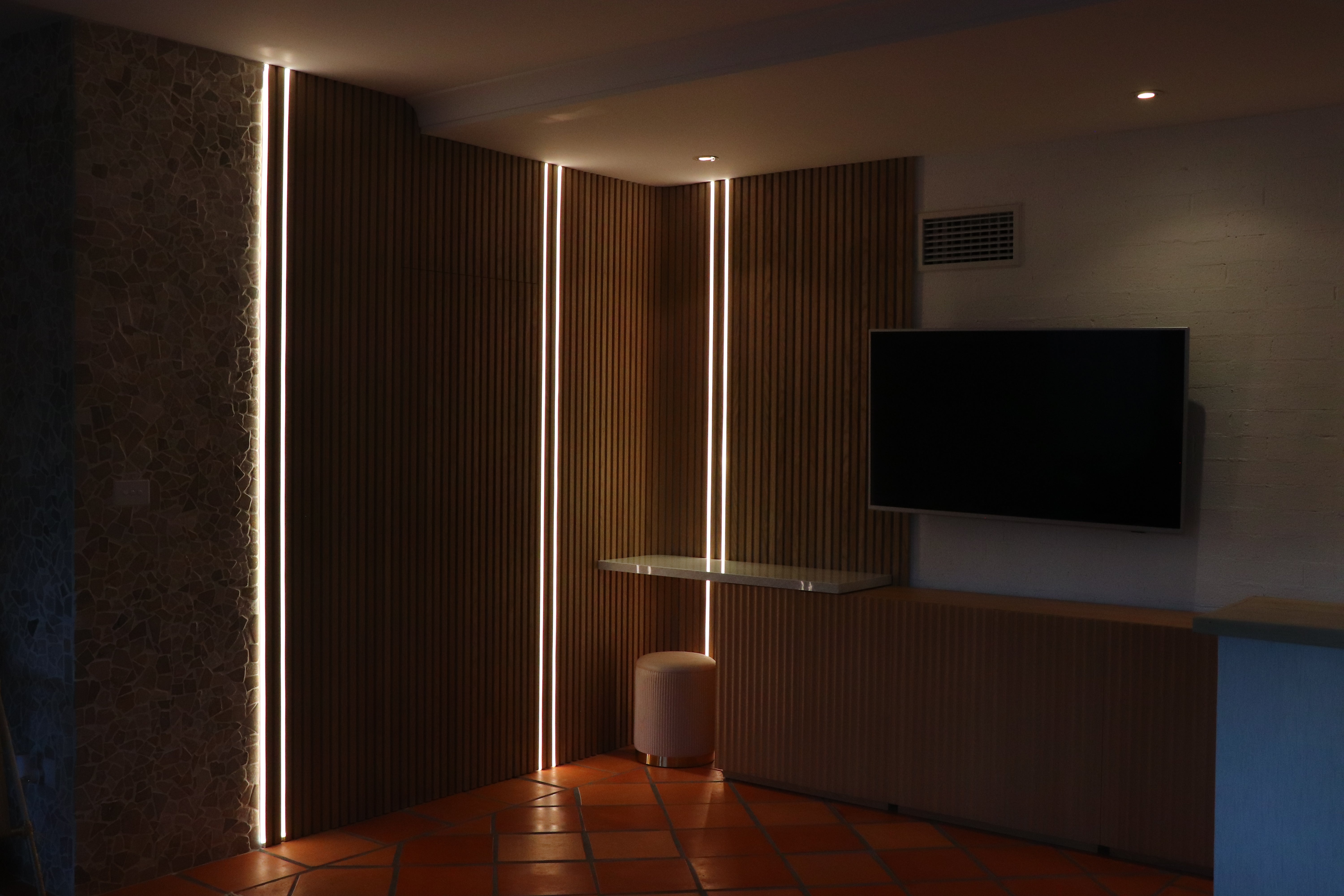 LED Lights recessed into wooden wall facade in a living room. Dimly lit room with a TV and downlights illuminating, creating a serene ambience.